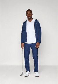 Casaco e calças desportivas azul-marinho com uma camisa polo branca. O conjunto apresenta linhas elegantes, e a pessoa segura um taco de golfe.