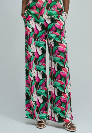 Pantalones de pierna ancha con vibrantes estampados de hojas en rosa, blanco y verde, combinados con tacones blancos de tiras y manos en los bolsillos sobre un fondo liso.