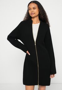 Μαύρο ριπ πλεκτό φόρεμα με χρυσό φερμουάρ, μακριά μανίκια και γιακά. Διαθέτει στενή εφαρμογή και τελειώνει πάνω από το γόνατο.