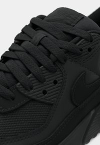Gros plan d'une chaussure de sport noire avec un mesh texturé, des lacets épais et arrondis, des surpiqûres et un logo Swoosh discret sur le côté.