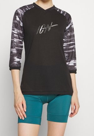 Person trägt ein schwarzes Sportshirt mit V-Ausschnitt, grauen Ärmeln mit Muster und glänzenden "IGN"-Buchstaben, kombiniert mit türkisfarbenen Radhosen.