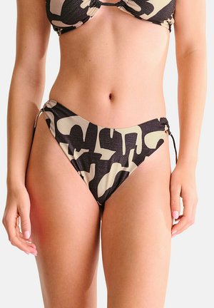 Femme portant un bas de bikini taille haute avec un motif camouflage abstrait noir et beige et des liens latéraux, montrée de la taille jusqu'au milieu des cuisses.