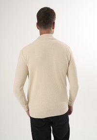 Cremefarbenes Strickpullover mit gerippter Struktur, Kragen und langen Ärmeln, getragen mit schwarzen Hosen, von hinten betrachtet.