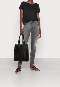 Bolso tote negro con textura de cocodrilo, forma rectangular y mangos dobles. Llevado con camiseta negra, jeans grises y sandalias negras.