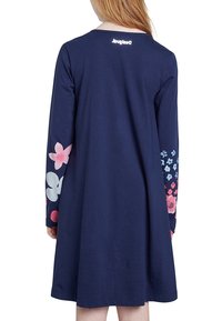 Femme portant une robe à manches longues bleu marine avec des motifs floraux colorés sur les manches, vue de dos, montrant le dos et le logo près de l'encolure.