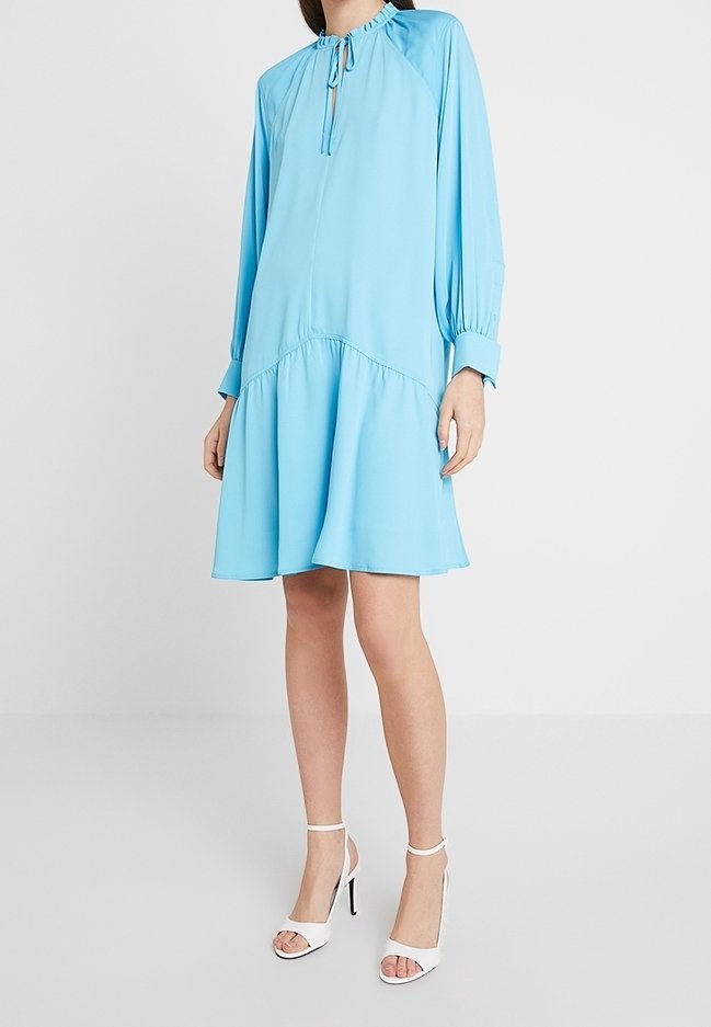 Femme portant une robe bleu clair ample, longueur genou, à manches longues, et des sandales blanches à talons hauts sur un fond uni.