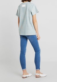 Koszula w kratę z krótkim rękawem w niebiesko-białe wzory z kwadratową metką na górnej części pleców, zestawiona z dopasowanymi niebieskimi dżinsami i białymi butami.