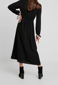 Schwarzes Langarmkleid mit geraffter Taille, fließendem Rock und gerafften Ärmeln mit Bindegurten. Getragen mit schwarzen, hohen Stiefeletten.