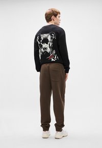 Μαύρο φούτερ με γραφικό σκύλου στην πλάτη και κόκκινο κείμενο "HUGO", συνδυασμένο με καφέ φόρμες και λευκά αθλητικά παπούτσια.