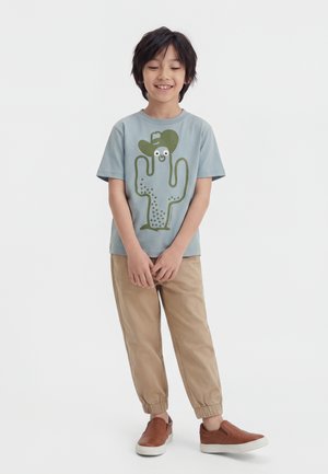 Ung dreng smiler, iført en blå t-shirt med en grøn kaktusgrafik, beige bukser og brune slip-on sko, står med hænderne foldet.