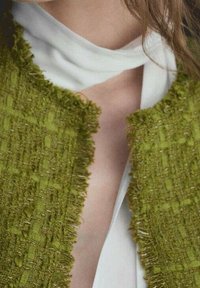 Chaqueta verde texturizada con bordes desgastados, superpuesta sobre una prenda blanca. El diseño presenta un corte sin cuello.