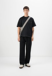 Svart t-skjorte med kort erme og lomme, kombinert med svarte vide bukser. Modellen har på seg en grå crossover-veske og hvite sneakers.