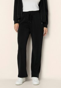 Osoba oblečená v černých teplákách se stahovací šňůrkou, bílém tričku, černé zipové bundě a bílých teniskách, stojící na jednotném pozadí.