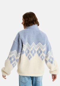 Pull en polaire bleu clair et crème avec un motif géométrique. Il présente un col haut, des manches longues et un tissu doux et texturé.