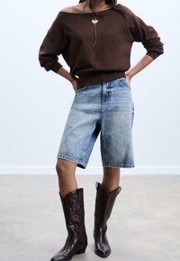Pull marron à manches tombantes, short en denim bleu clair légèrement délavé, et bottes de cowboy marron avec des motifs embossés.