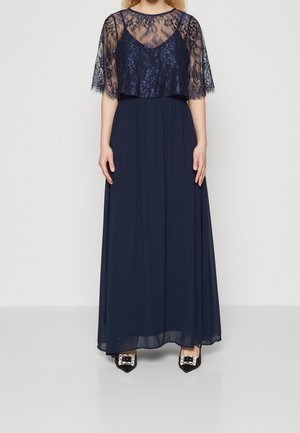 Kobieta w granatowej sukni do ziemi z koronkowymi krótkimi rękawami oraz zdobionych czarnych czółenkach na szpic, stojąca na białym tle.