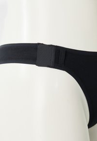 Crna tanga donji veš s glatkom teksturom, koji ima široku elastičnu traku i minimalistički dizajn. Bešavne ivice za udobnost.