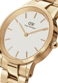 Reloj de tono dorado con caja redonda, esfera blanca, marcadores de horas minimalistas y pulsera de metal enlazada. Nombre de la marca "Daniel Wellington" mostrado.