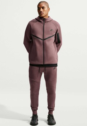 Mężczyzna w pasującej mauve bluzie z kapturem Nike i spodniach dresowych oraz czarnych sneakersach stoi na jednolitym jasnym tle.