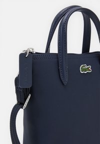 Sac à main synthétique bleu marine avec deux poignées supérieures, fermeture éclair et petit patch logo Lacoste. Comprend une bandoulière amovible et une surface texturée.