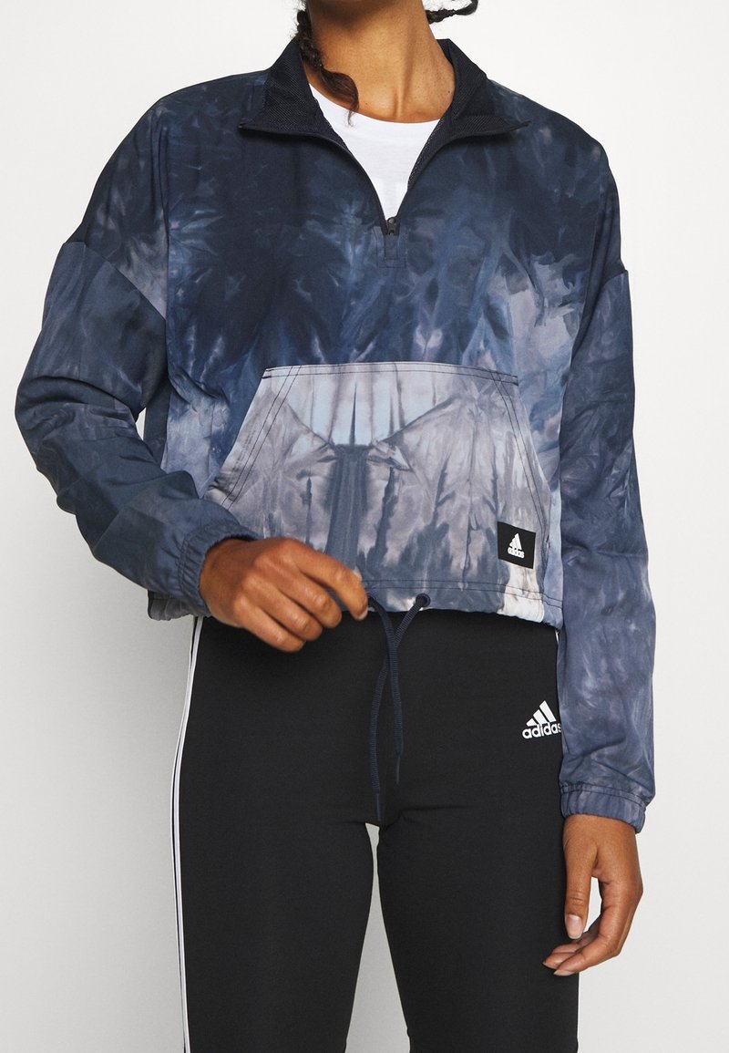 Personne portant une veste tie-dye bleu marine et grise à demi-fermeture éclair avec poche avant et des leggings noirs Adidas avec logo blanc et bandes latérales.