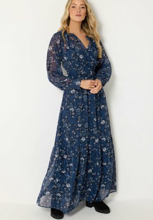 Donna che indossa un lungo vestito floreale blu navy con maniche lunghe di tulle e stivali neri, in piedi davanti a uno sfondo chiaro e semplice.