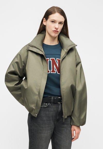 Νεαρή γυναίκα με ίσια καστανά μαλλιά, ντυμένη με χακί μπουφάν πάνω από μπλε φούτερ με κόκκινα και λευκά γράμματα, συνδυασμένο με μαύρο τζιν.