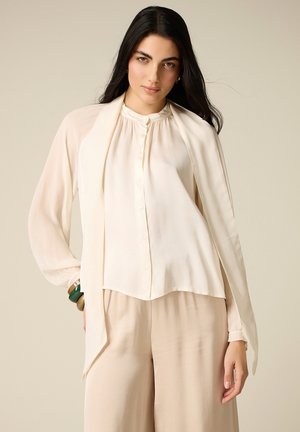 Giovane donna con lunghi capelli scuri indossa una camicetta crema con dettaglio a sciarpa, pantaloni beige a gamba larga e braccialetti verdi e marroni, posando con calma.