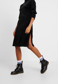 Svart långärmad klänning med hög krage och sidslitsar, kombinerad med svarta snörboots och vita strumpor med blå ränder.