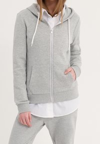 Sudadera con cremallera de color gris claro, fabricada en un tejido suave, con capucha ajustable, bolsillos tipo canguro y puños de canalé, superpuesta sobre una camiseta blanca.