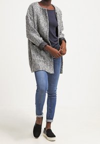 Cardigan long texturé gris et blanc porté sur un haut bleu marine, associé à un jean skinny bleu clair et des chaussures noires sans lacets.