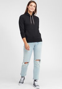 Schwarzer Hoodie aus Baumwolle mit hohem Kragen und verstellbaren Kordeln. Kombiniert mit hellblauen, ausgewaschenen Jeans und schwarzen Sneakers.