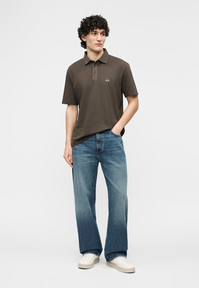 Jeune homme debout, main dans la poche, portant un polo marron, un jean large bleu et des baskets blanches, regardant sur le côté.