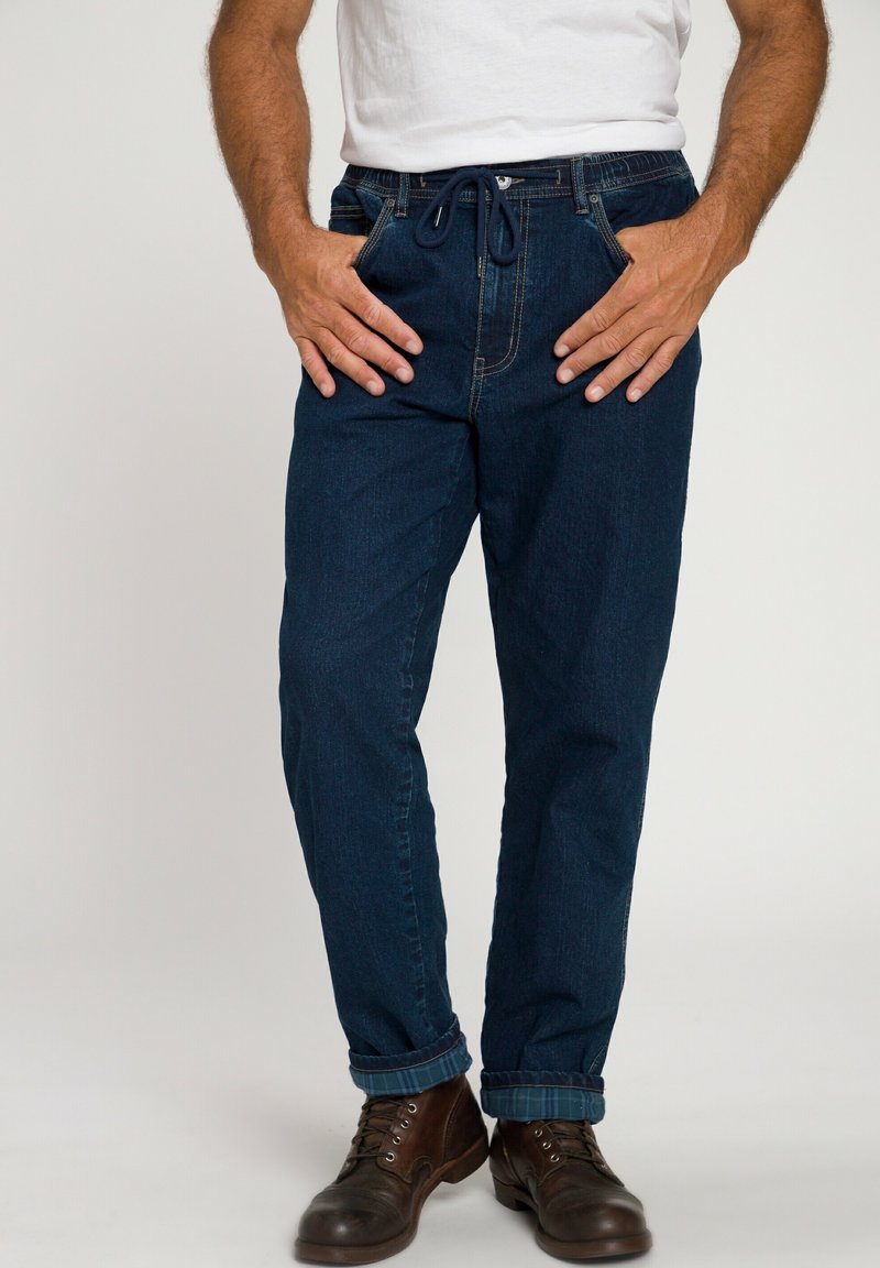 Pantalones de mezclilla azul oscuro con cintura elástica y cordón, piernas ajustadas y puños remangados, combinados con botas marrones de cordones.