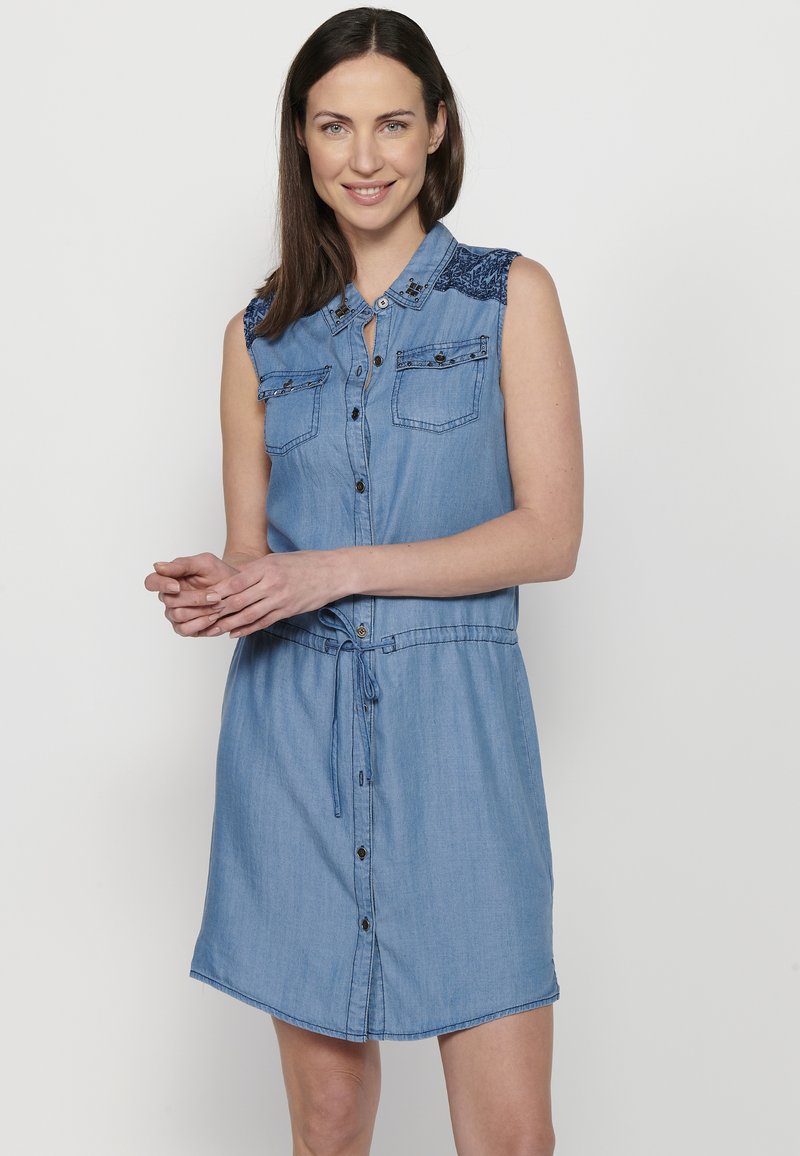 Abito in denim con vita definita da una cintura, maniche corte, tasche sul petto e dettagli ricamati sulle spalle. Colore blu chiaro generale.