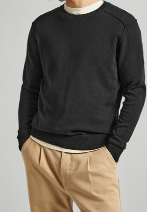 Mann in schwarzem Rundhalspullover über weißem Hemd, kombiniert mit beigen, gefalteten Hosen, Hände in den Taschen, vor einem schlichten Hintergrund.