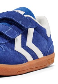 Blå mockasneakers med vita ränder, kardborreband och en texturerad orange gummisula. Närbild av design och material.