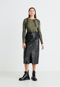 Haut à manches longues vert olive avec une texture côtelée, associé à une jupe midi en simili cuir noire avec une fente avant et un détail de ceinture. Bottes noires à lacets.