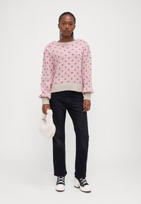 Lys pink sweater med mørkere pink prikker, ribbet halsudskæring og manchetter. Sammen med sorte jeans og hvide sko, holder en cremefarvet håndtaske.