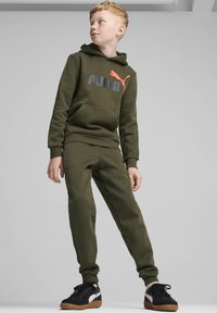 Conjunto de sudadera y pantalones de chándal color verde oliva con un bolsillo tipo canguro, que presenta un logo de Puma en gris y naranja en la parte frontal. Zapatillas negras complementan el look.