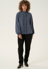 Blusa grigia a balze con colletto alto, chiusura con bottoni frontale e maniche lunghe, abbinata a pantaloni neri e stivali alla caviglia con punta affilata.