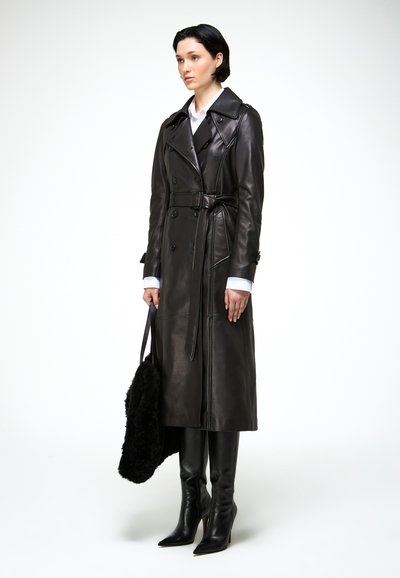 Femme portant un long trench-coat en cuir noir, des bottes noires montant jusqu'aux genoux, tenant un sac fourrure noir, se tenant devant un fond blanc uni.
