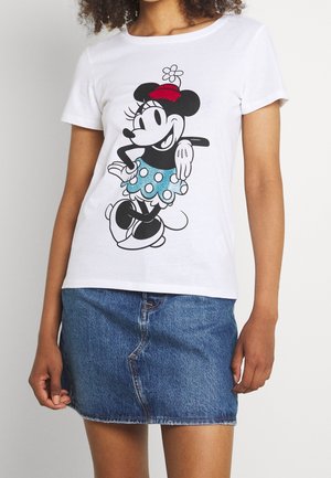 Osoba ubrana w białą koszulkę z kolorową grafiką Minnie Mouse oraz niebieską spódnicę dżinsową, stojąca na tle jednolitego jasnego tła.