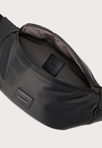 Fekete nylon deréktáska cipzáras nyitással, sima textúrával és dombornyomott logóval. Belseje szürke anyagból készült, tágas kialakítással.