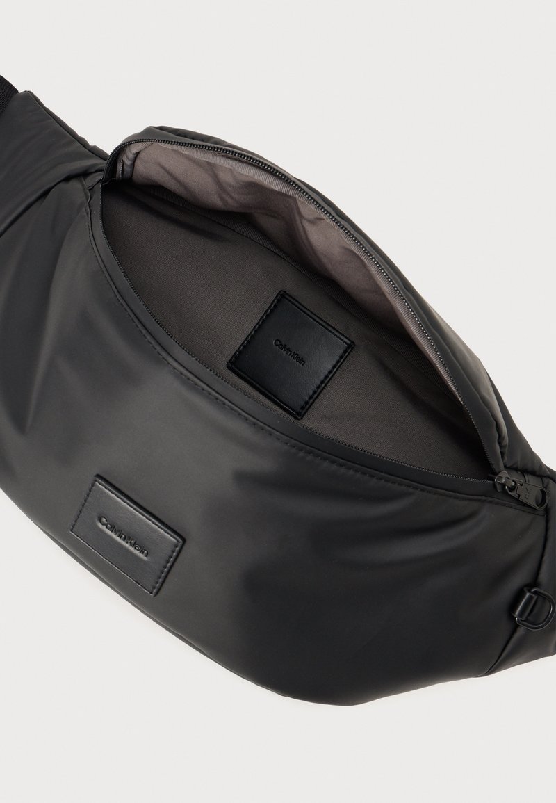 Sac banane en nylon noir avec une ouverture à fermeture éclair, texture lisse et patch logo en relief. Intérieur en tissu gris, design spacieux.