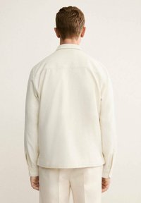 Camisa de manga larga color beige claro, hecha de una tela suave, con un collar clásico y detalles de costura sutiles en la parte de atrás.