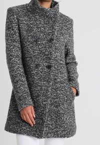 Abrigo de lana texturizada gris y negro con un alto cuello, delante de doble botonadura y dos bolsillos laterales. Cuenta con botones negros y un corte ajustado.