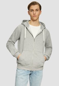 Grijze zip-up hoodie met een trekkoordcapuchon, voorzien van voorzakken en geribbelde manchetten, gedragen over een wit t-shirt, gecombineerd met lichtblauwe spijkerbroek.