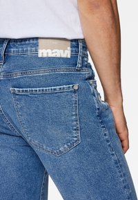 Blå denimjeans med en kroppsnära design, försedd med en bakficka och en etikett på midjebandet. Materialet har en textur med synliga sömmar.