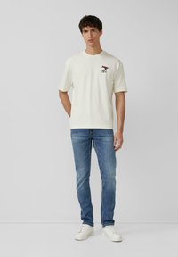 Junger Mann mit einem weißen T-Shirt, auf dem ein kleiner roter Cartoon-Skifahrer auf der Brust abgebildet ist, blauen Jeans und weißen Turnschuhen, der vor einem schlichten hellen Hintergrund steht.